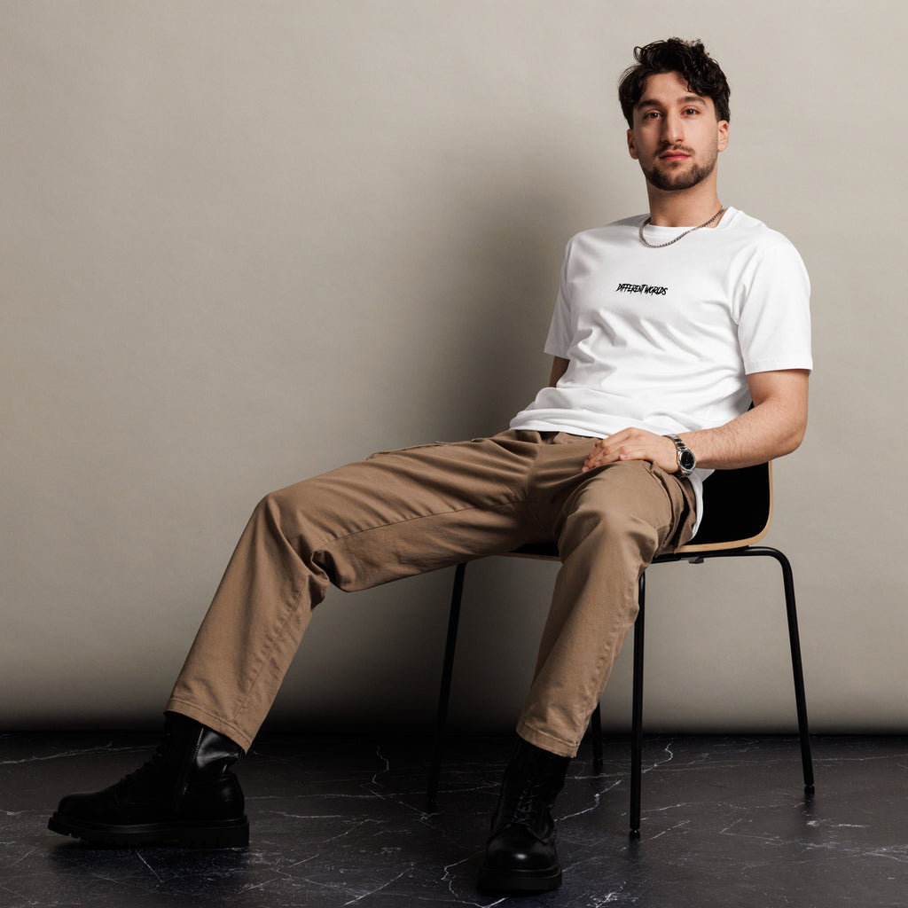 Man sitting on a chair wearing a white "Different worlds" t-shirt and beige pants against a plain background