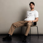 Man sitting on a chair wearing a white "Different worlds" t-shirt and beige pants against a plain background
