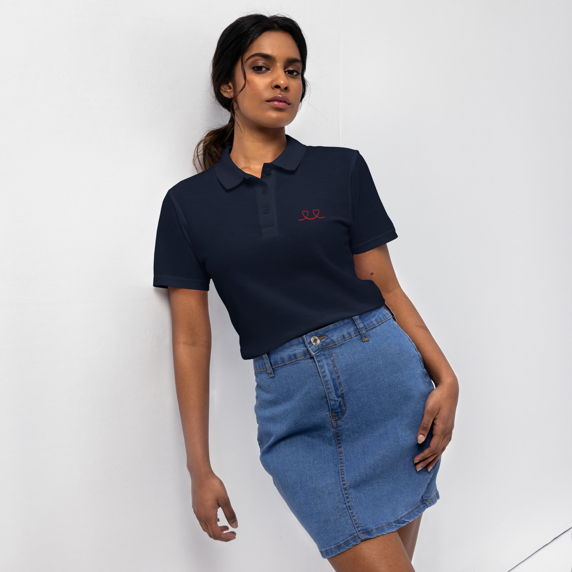 Woman wearing a navy polo shirt with a red love smiley embroidery and a blue denim skirt on a white background