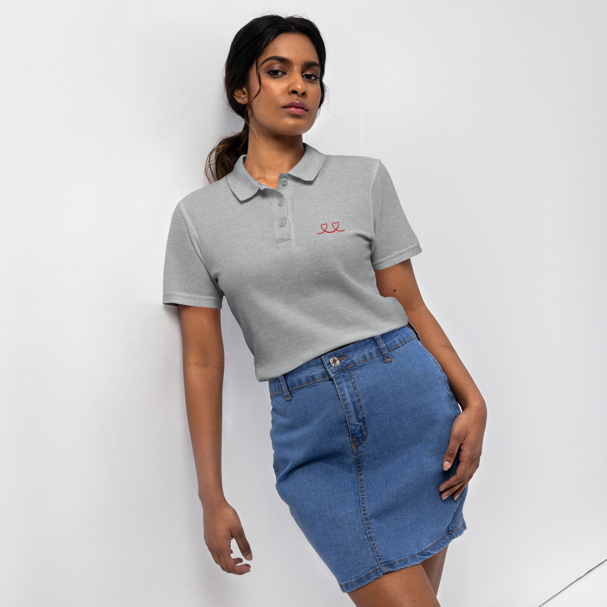 Woman wearing a sport grey polo shirt with a red love smiley embroidery and a blue denim skirt on a white background