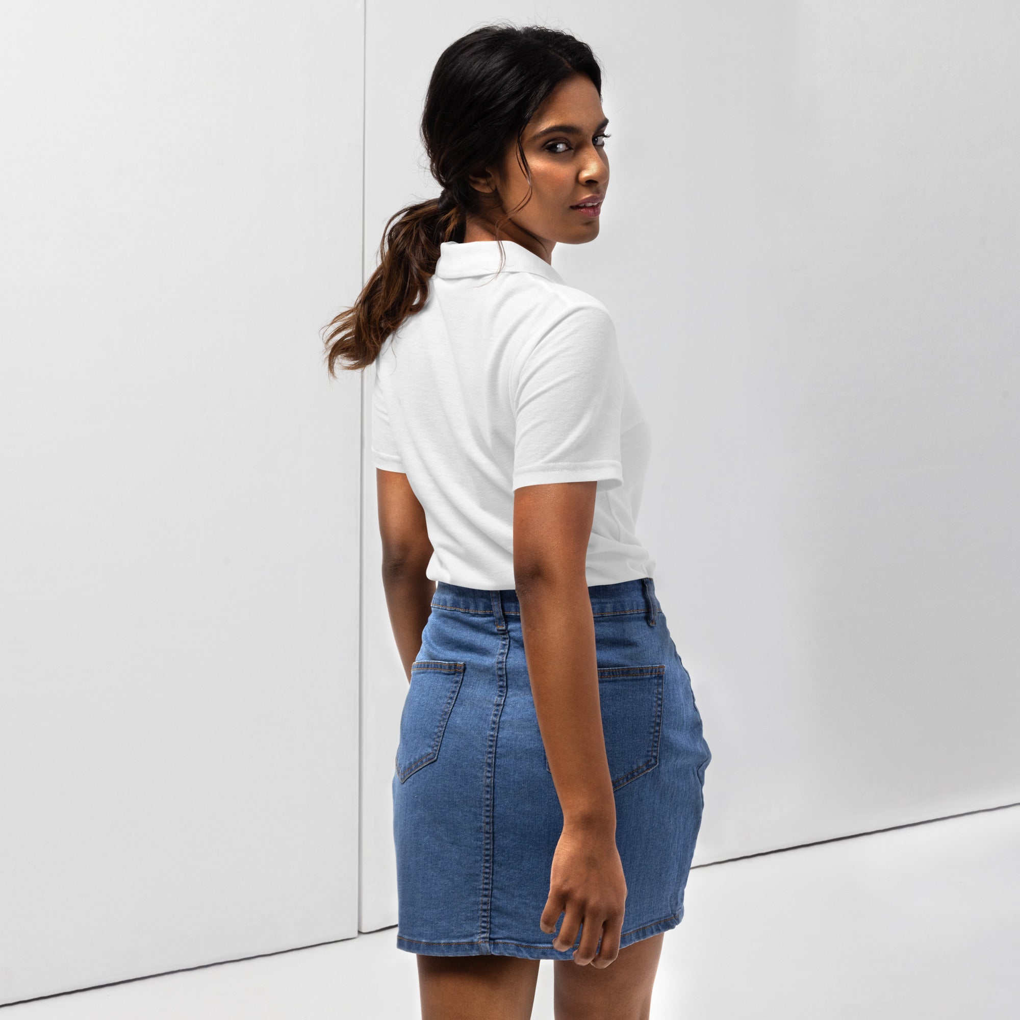 Woman wearing a white pique polo shirt with a red love smiley embroidery and a blue denim skirt on a white background. Photo taken from the backside.