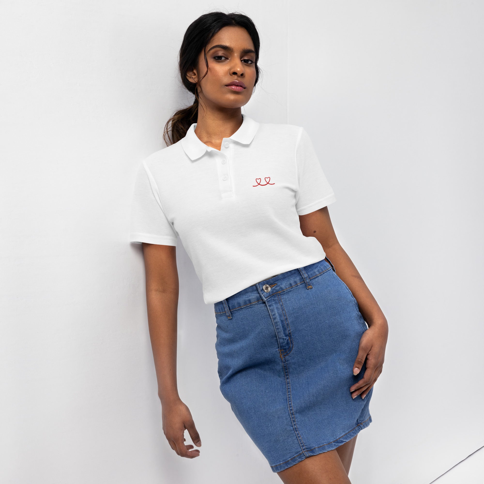 Woman wearing a white pique polo shirt with a red love smiley embroidery and a blue denim skirt on a white background