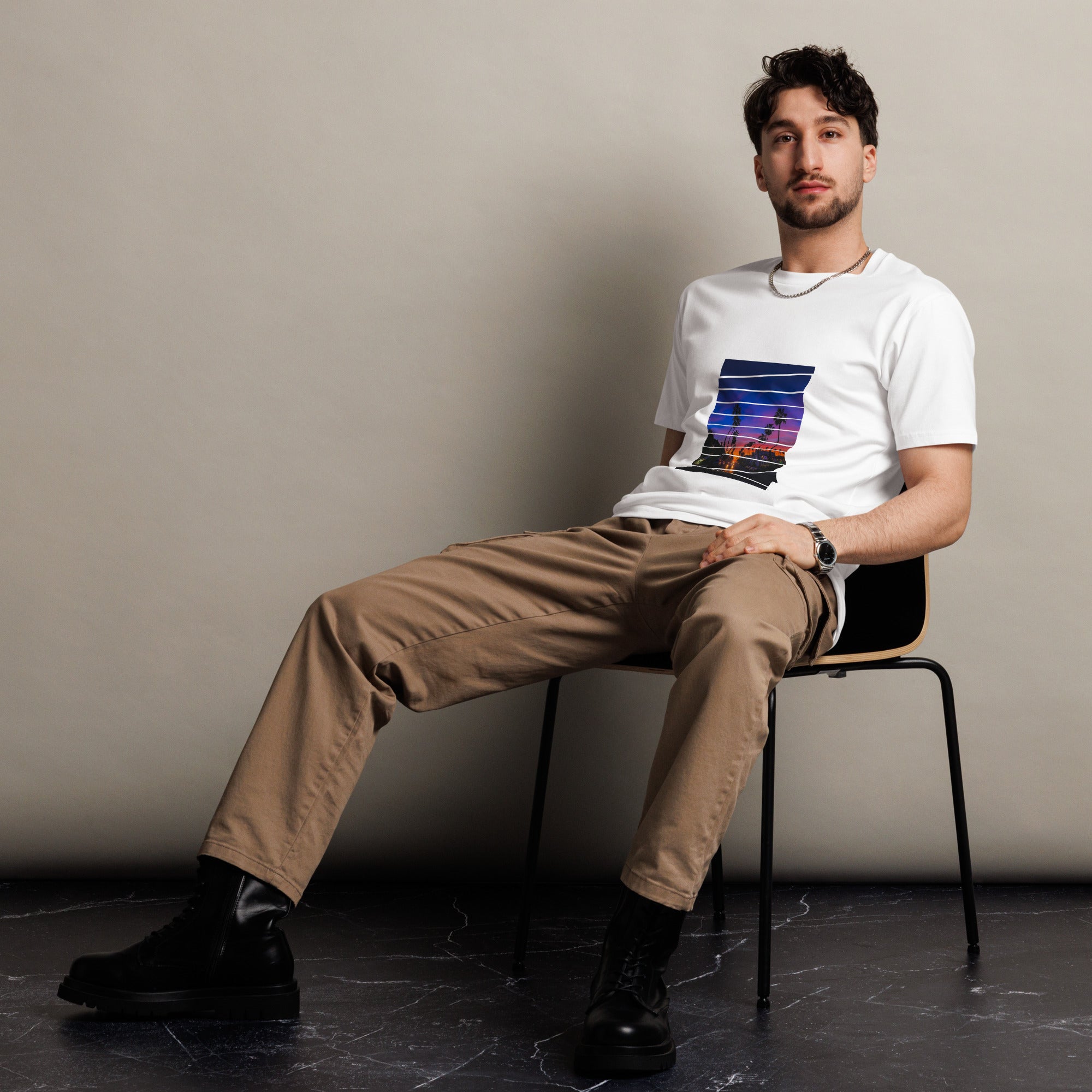Man sitting on a chair wearing a white t-shirt with a colorful sunset graphic and beige pants.